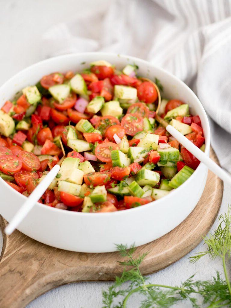 Insalata di Cetrioli, Pomodori, Cipolle e Avocado 2 Ciotola bianca con insalata di cetrioli e pomodori e pinze da insalata appoggiate all'interno, su un tagliere di legno.