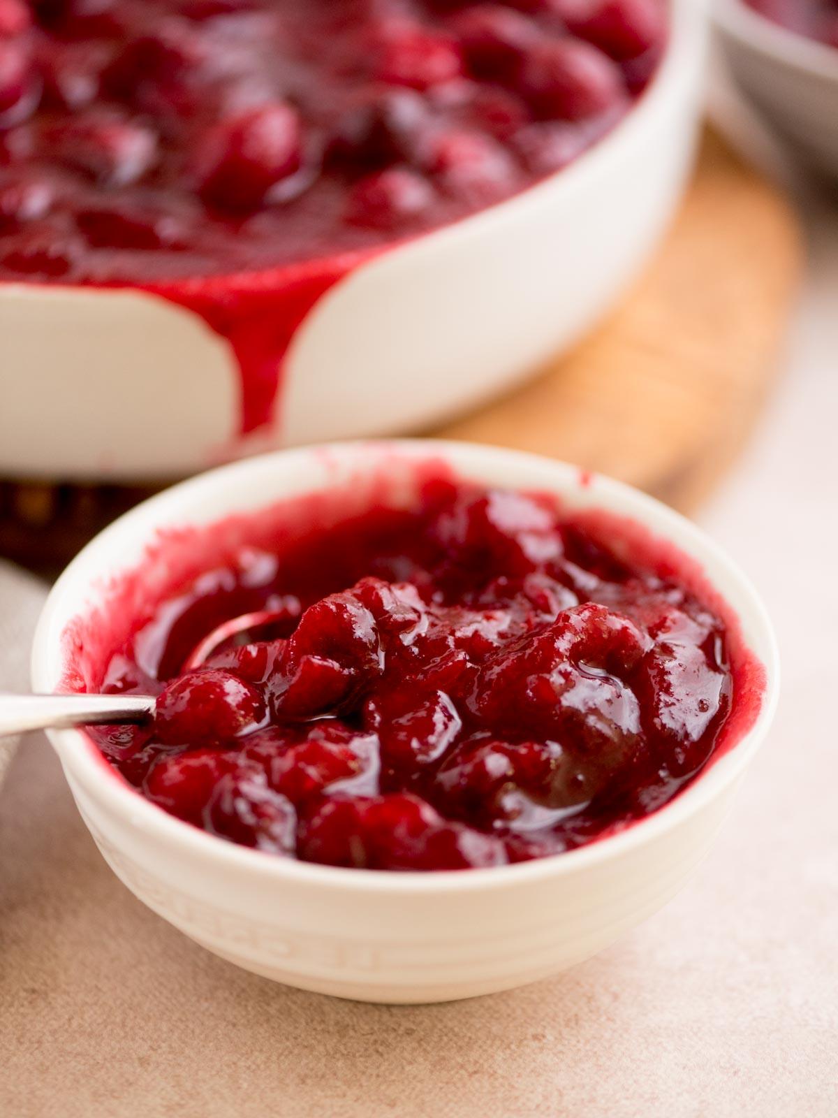 Salsa ai Mirtilli Rossi Interi 2 Una vista ravvicinata di una piccola ciotola bianca piena di salsa di mirtilli rossi fatta in casa, che mostra mirtilli interi lucidi. Una ciotola più grande con più salsa è sullo sfondo, leggermente traboccante. Un cucchiaio è nella piccola ciotola.