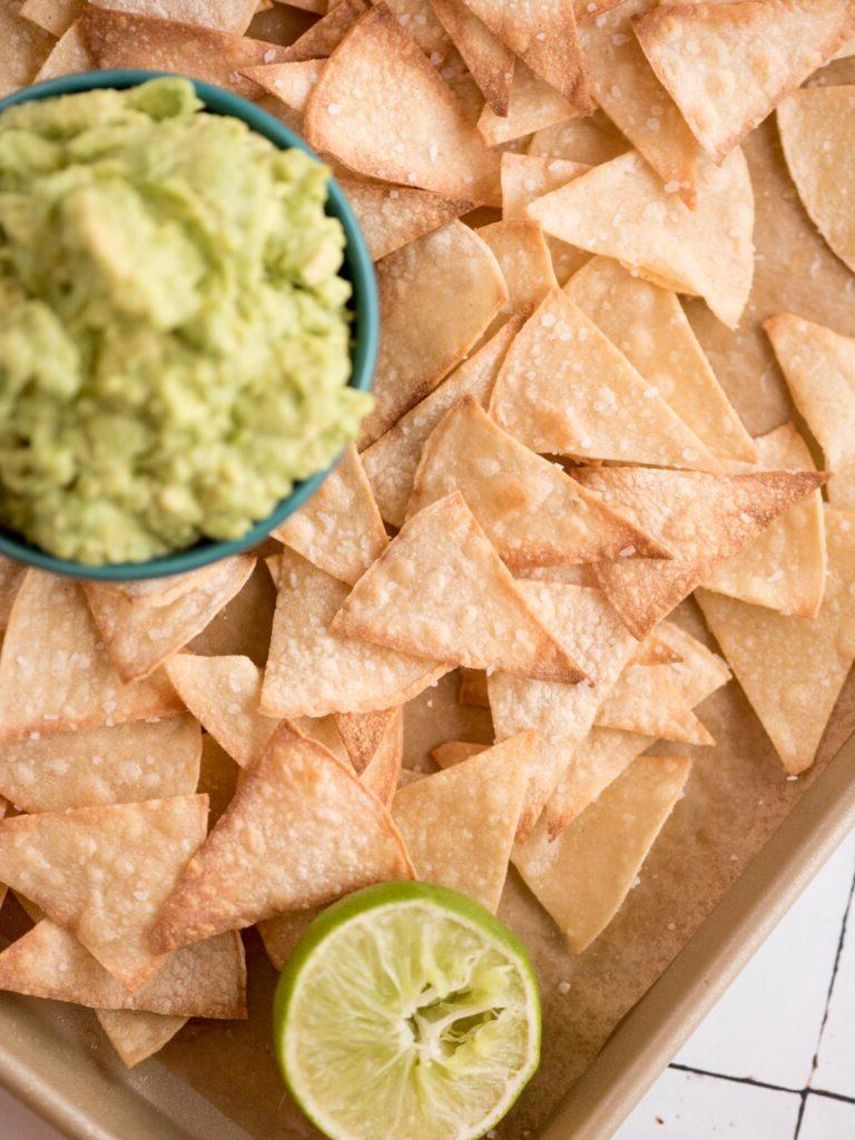 Una piccola ciotola di guacamole incastonata in un vassoio di chips di tortilla al forno con mezzo lime spremuto.