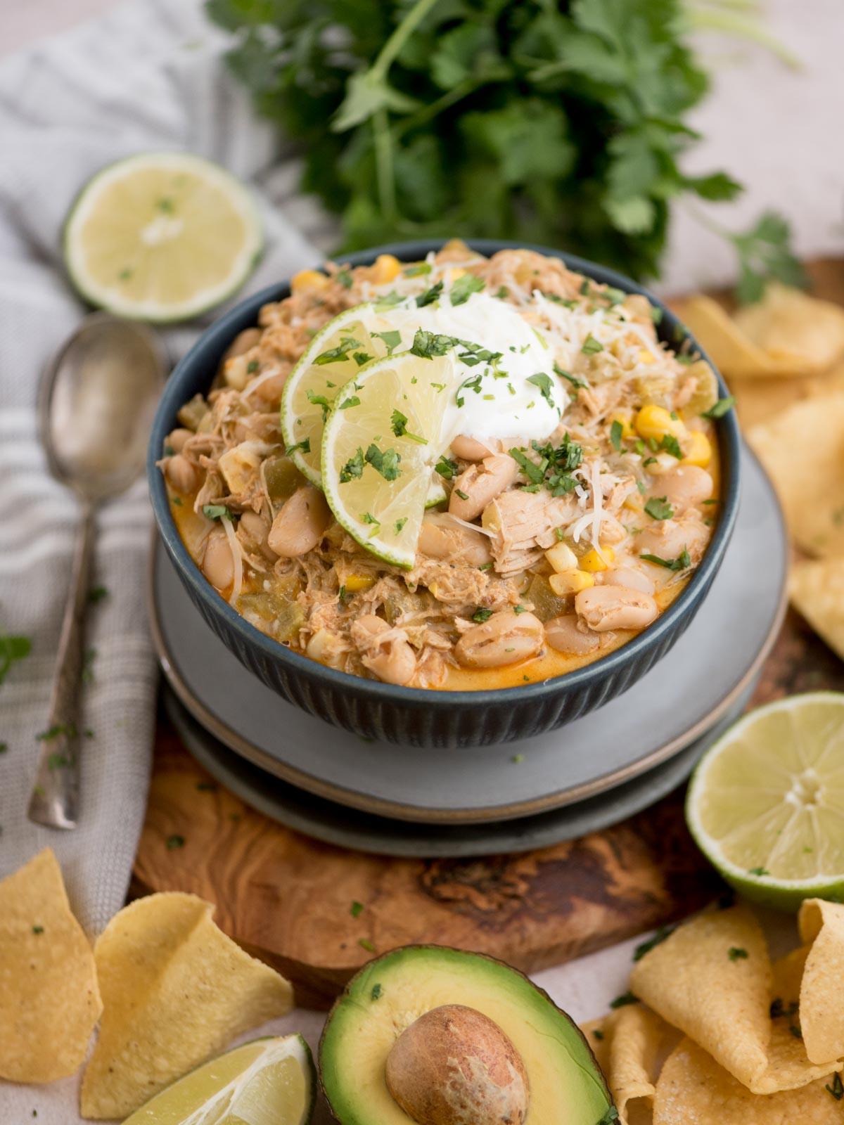 Chili di Pollo Bianco del Sud-Ovest 2 Una ciotola di chili bianco di pollo guarnito con fette di lime, panna acida e coriandolo. Ingredienti come avocado, tortilla chips e mezze lime circondano la ciotola. Un cucchiaio e un mazzetto di coriandolo sono posizionati nelle vicinanze.