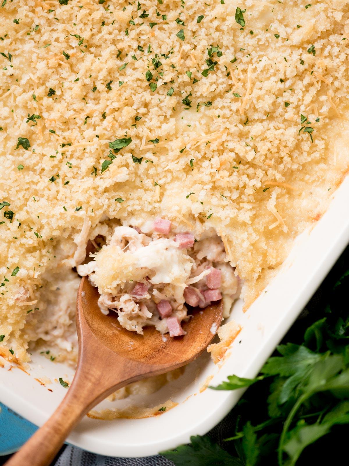 Una teglia di pollo cordon bleu pronta da servire, con un cucchiaio da portata inserito. Attorno, prezzemolo fresco e un canovaccio da cucina.