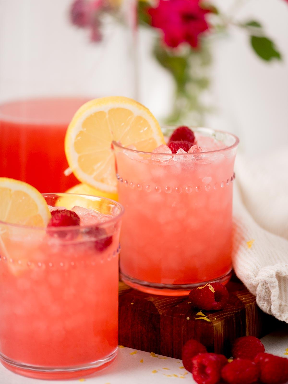 Due bicchieri di limonata rosa ai lamponi con ghiaccio, guarniti con fette di limone e lamponi, posati su un tagliere di legno. Lamponi freschi e un panno bianco sono vicini, con fiori sfocati sullo sfondo.