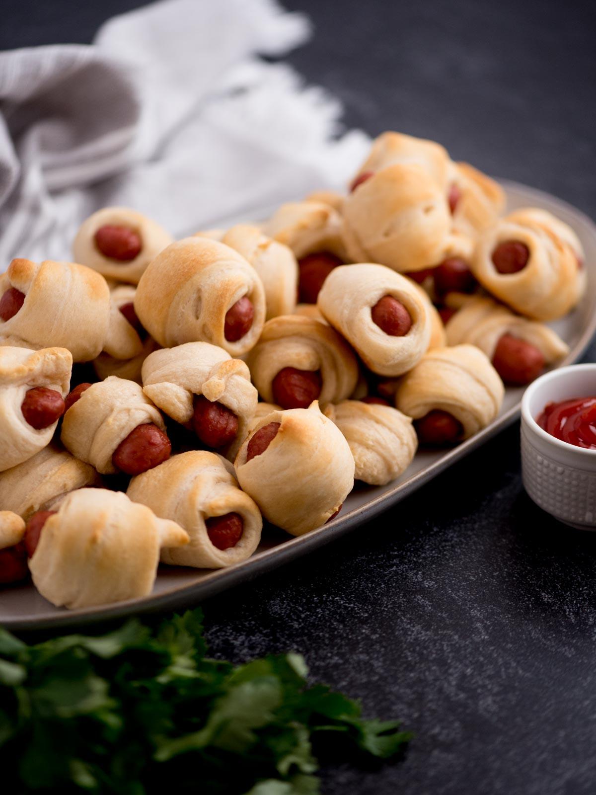 Mini Wurstel in Pasta Sfoglia 2 Pigs in a blanket, ovvero mini wurstel in sfoglia, impilati su un piatto con una piccola ciotola di ketchup.