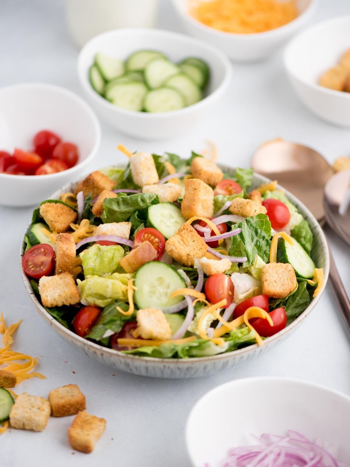 Ricetta Insalata della Casa 2 Insalata di casa classica in una ciotola, circondata da utensili da portata e condimenti freschi.