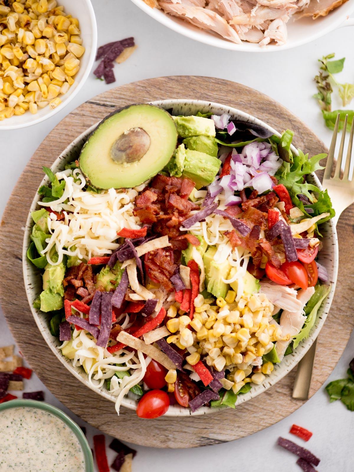 Insalata di Pollo del Sud-Ovest 2 Una colorata insalata in una ciotola con lattuga, avocado a fette, mais, formaggio grattugiato, pomodorini, cipolla rossa, pancetta croccante e strisce di tortilla multicolore. Una forchetta è appoggiata accanto, con ingredienti come mais e salsa in piccole ciotole vicine.