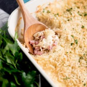 Una teglia di pollo cordon bleu pronta da servire, con un cucchiaio da portata inserito. Attorno, prezzemolo fresco e un canovaccio da cucina.