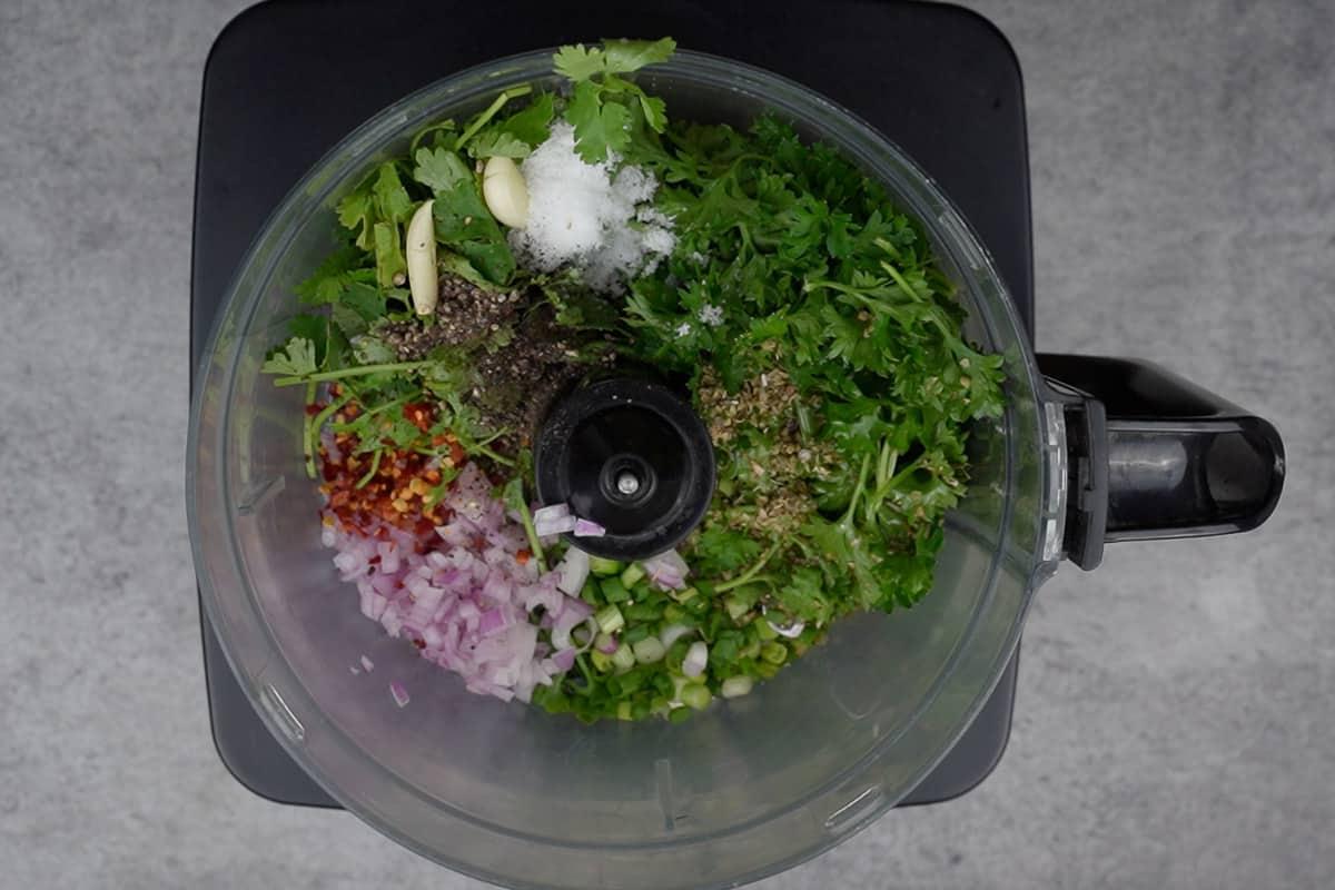 Coriandolo, prezzemolo, origano, cipollotti, cipolla rossa, aglio, sale, pepe e fiocchi di peperoncino aggiunti in un robot da cucina.