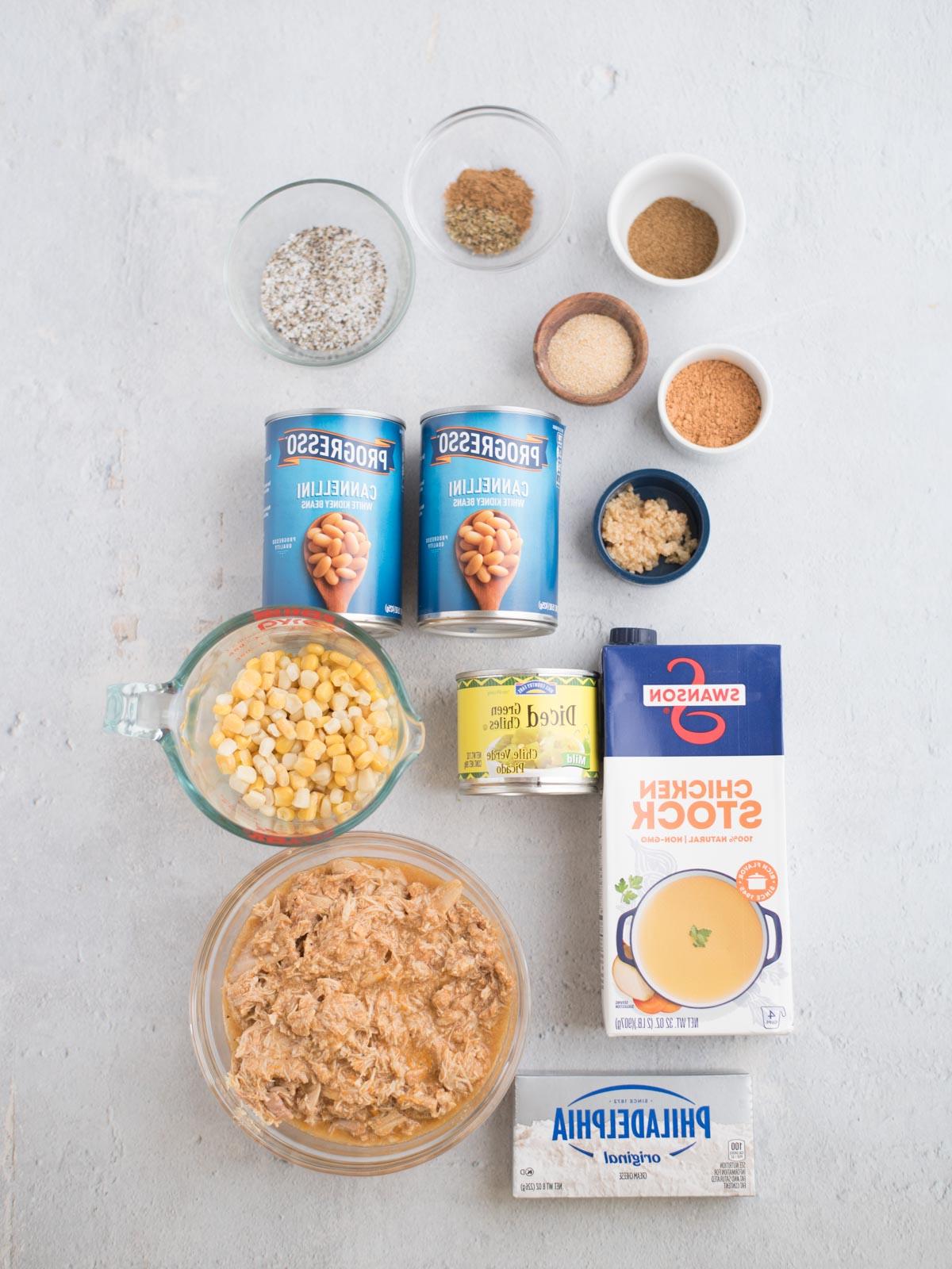 Chili di Pollo Bianco del Sud-Ovest 3 Ingredienti per un chili bianco di pollo del sud disposti artisticamente sulla superficie, con brodo di pollo, formaggio cremoso, fagioli cannellini in scatola, peperoncini verdi a cubetti, mais, pollo sfilacciato e diverse piccole ciotole di spezie e condimenti.