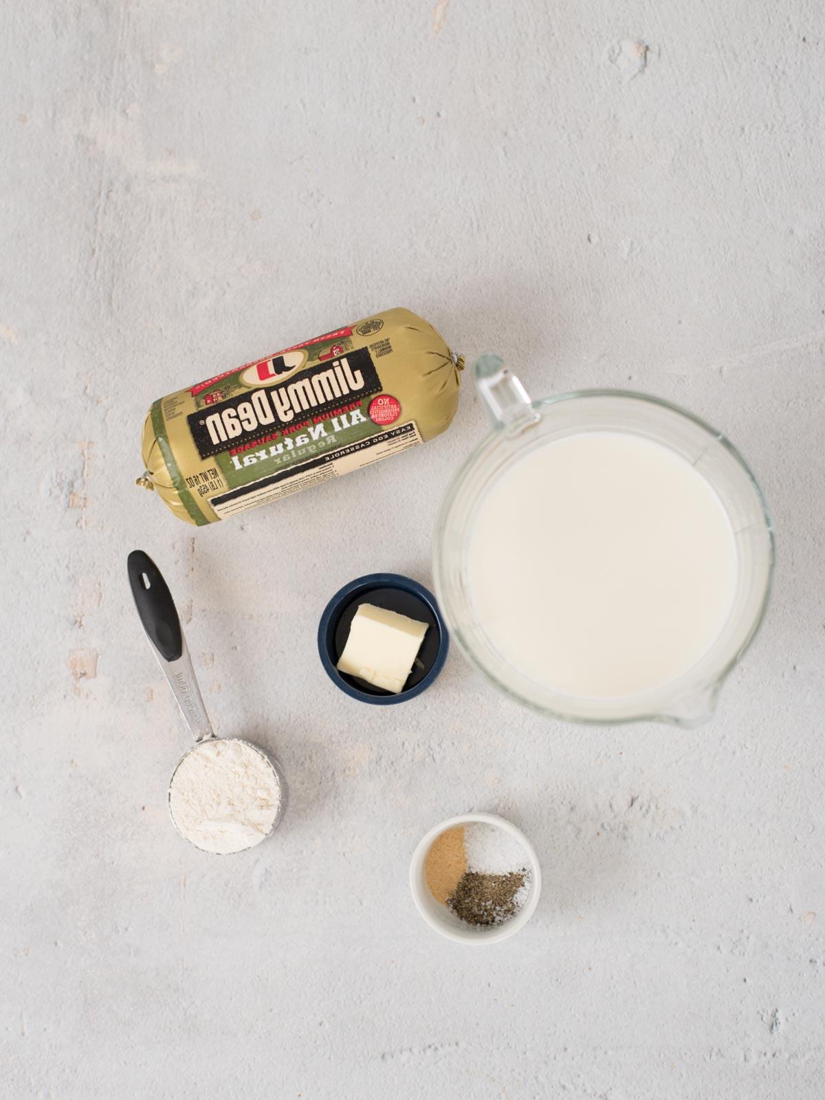 Sugo di Salsiccia 3 Una vista dall'alto degli ingredienti della salsa gravy per la colazione su una superficie chiara: una confezione di salsiccia Jimmy Dean, una caraffa di latte, una piccola ciotola con burro, una tazza dosatrice con farina e un piccolo piatto contenente sale e pepe.