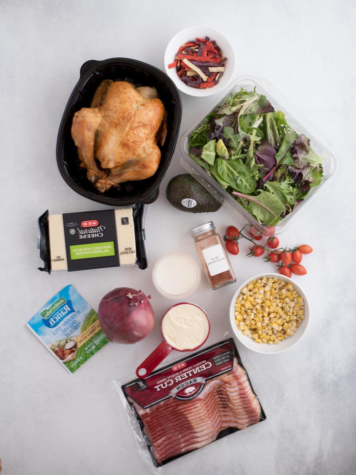 Insalata di Pollo del Sud-Ovest 3 Una disposizione a vista di vari alimenti, inclusi un contenitore di insalata mista, un pollo intero cotto, pomodorini ciliegino, un avocado, un blocco di formaggio, condimento per insalata, cipolla rossa, spezie, mais, condimento Ranch e una confezione di pancetta su una superficie chiara.