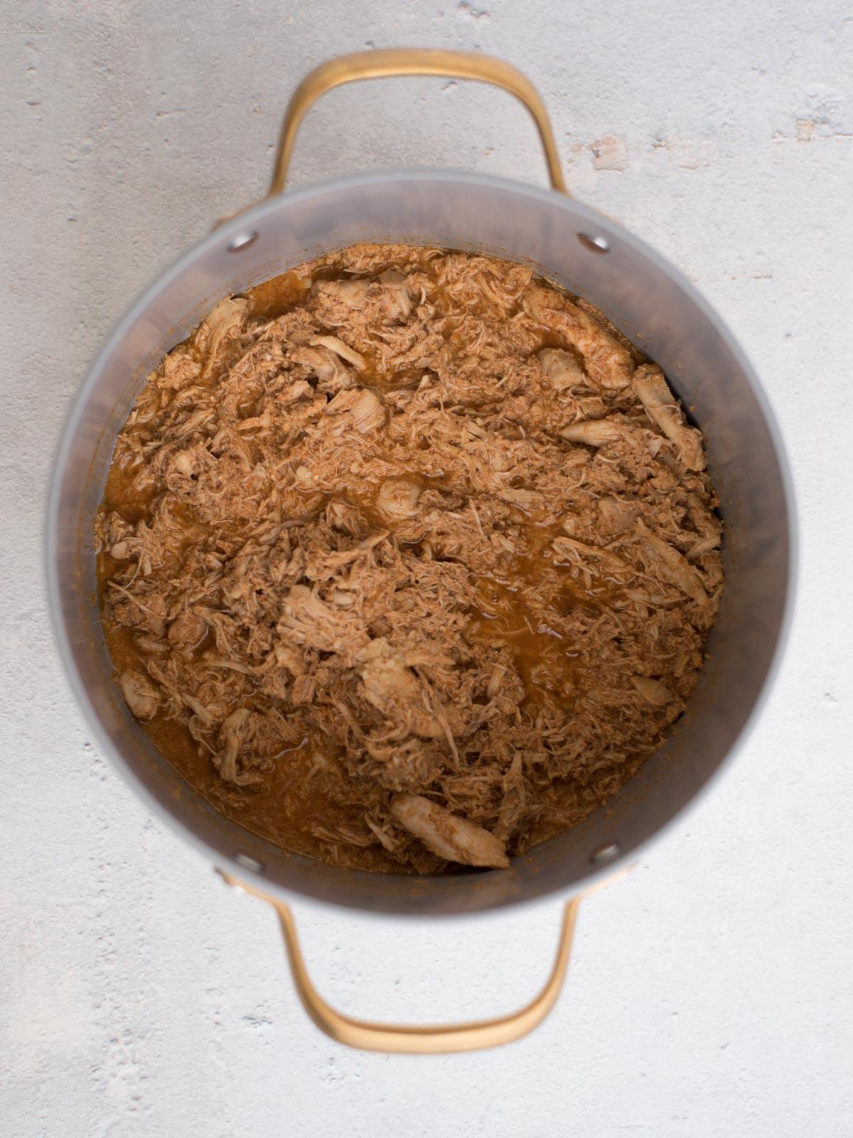Chili di Pollo Bianco del Sud-Ovest 4 Vista dall'alto di una grande pentola di metallo piena di carne sfilacciata tenera e succosa in una salsa marrone per un chili bianco facile con pollo. La pentola, completa di manici su entrambi i lati, poggia su una superficie chiara.