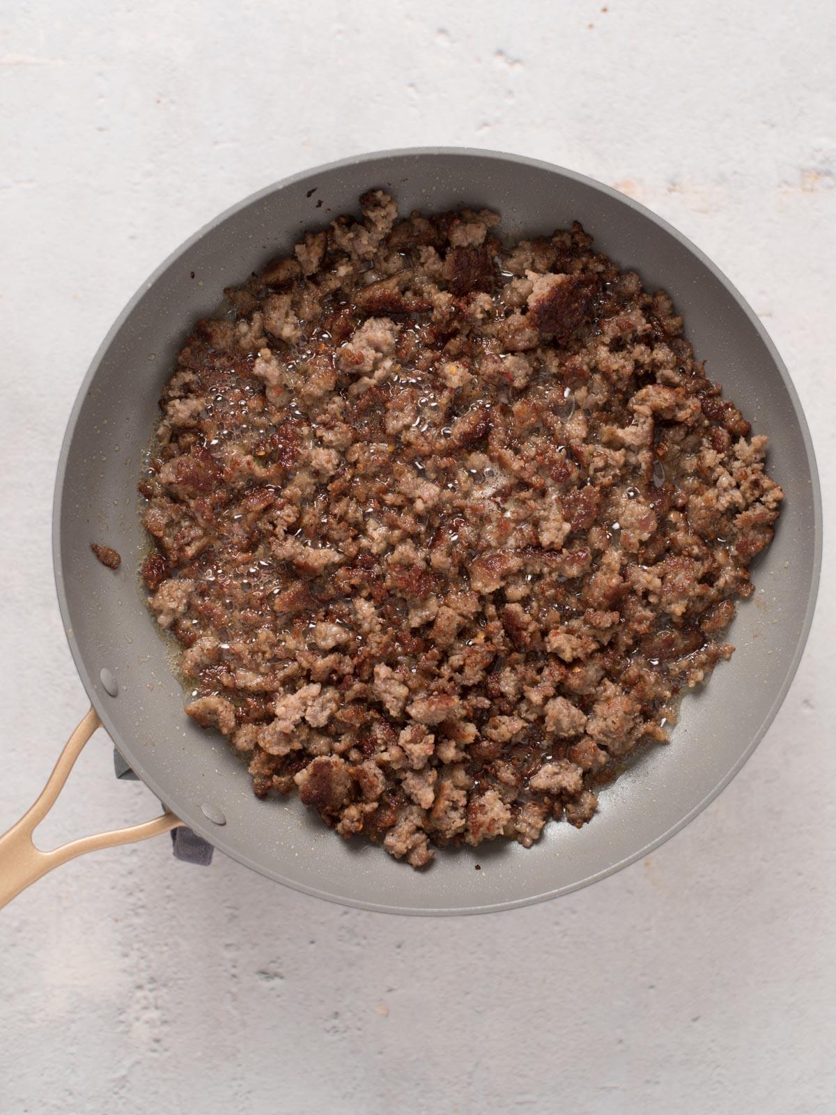 Sugo di Salsiccia 4 Una padella contenente carne macinata rosolata e sbriciolata è posizionata su una superficie chiara. La carne è cotta uniformemente, con una consistenza leggermente croccante. La padella ha un interno grigio chiaro e un manico in legno.
