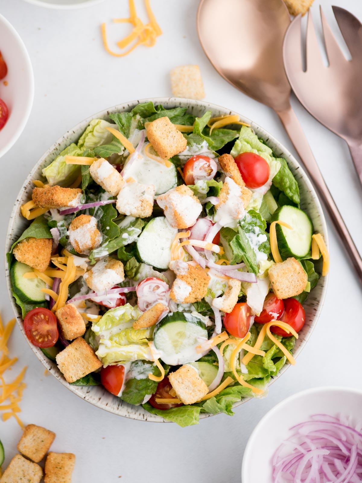 Ricetta Insalata della Casa 4 Insalata di casa in una ciotola, generosamente condita con salsa ranch, circondata da utensili da portata e vari topping freschi.