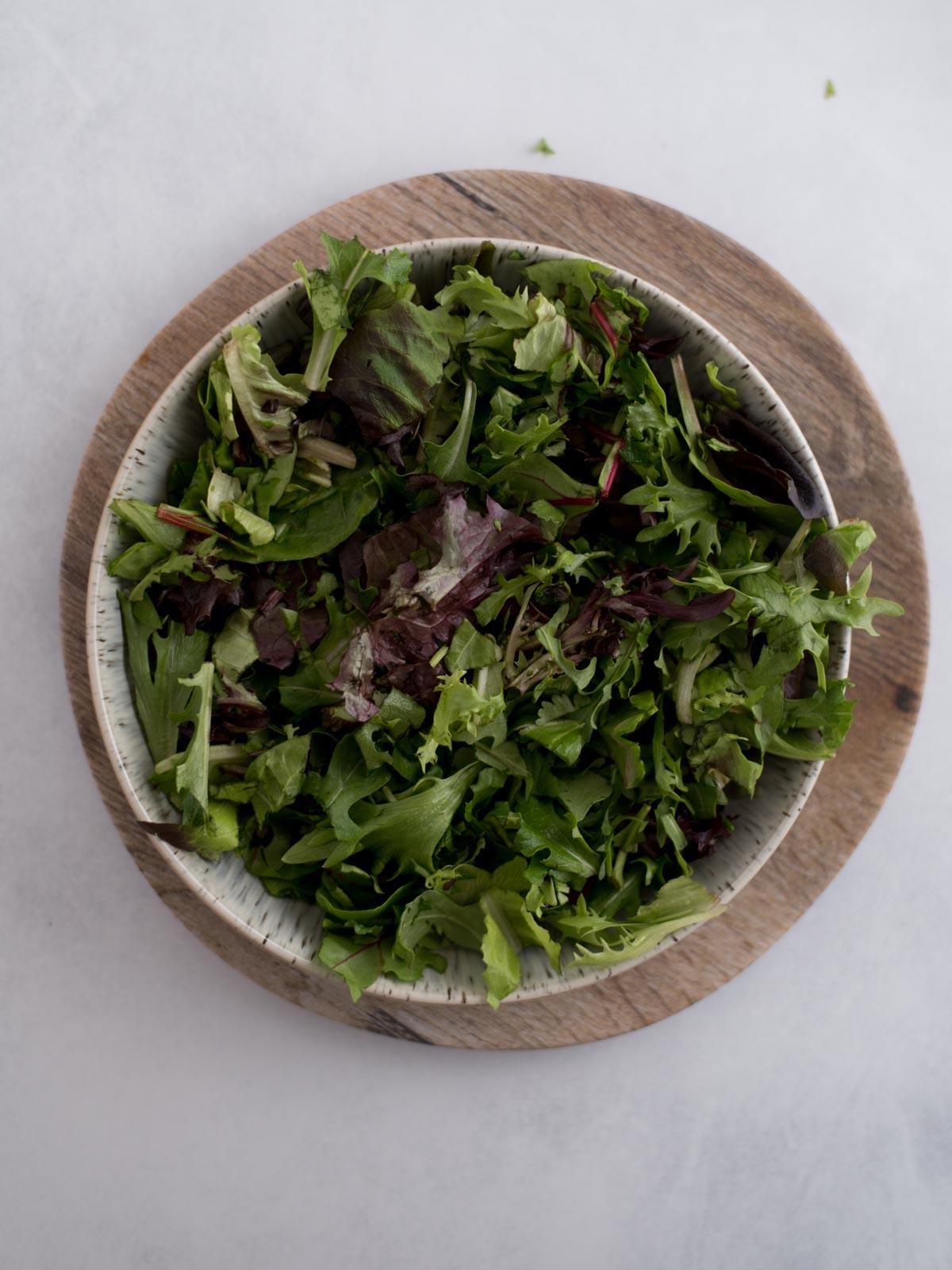 Insalata di Pollo del Sud-Ovest 4 Una ciotola di insalata a foglie miste si trova su una tavola di legno. Le verdure variano nelle tonalità di verde e viola, con diverse texture e forme, creando un aspetto fresco e vibrante su uno sfondo chiaro.
