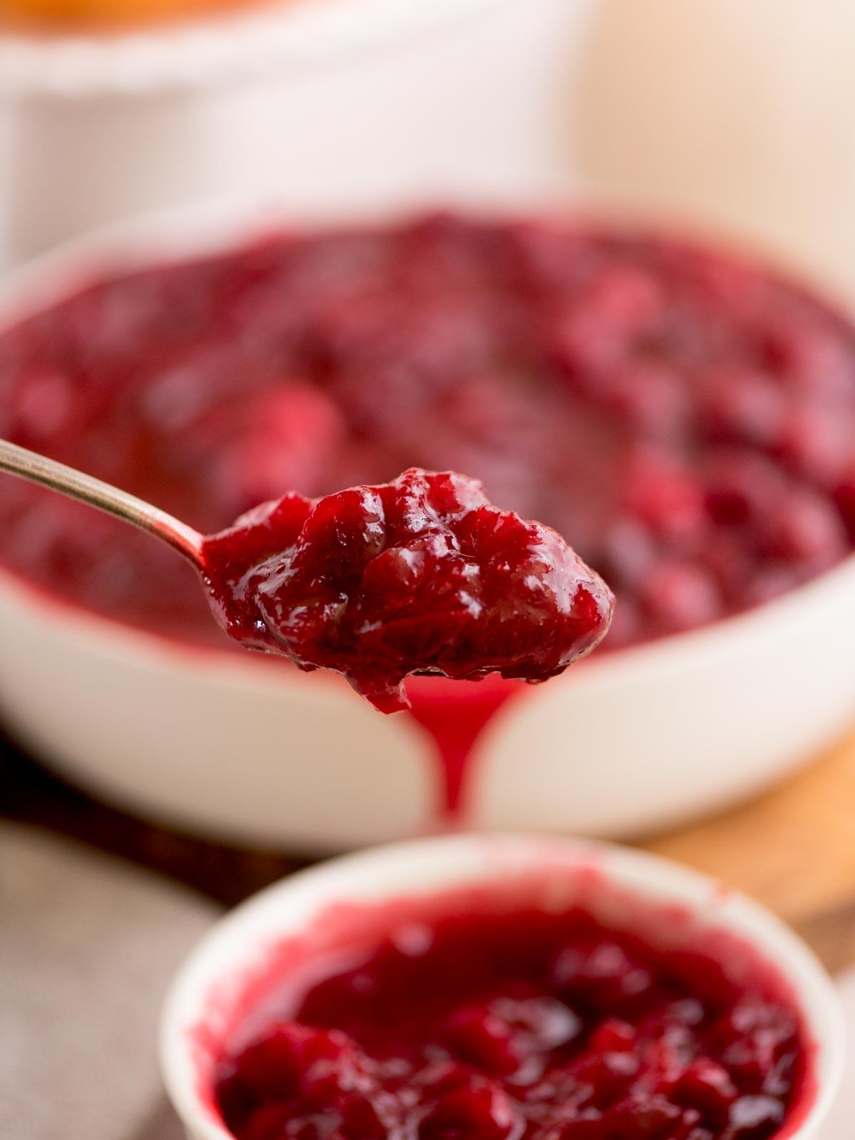 Salsa ai Mirtilli Rossi Interi 5 Un cucchiaio di salsa di mirtilli rossi è tenuto sopra una ciotola piena della stessa salsa. La salsa di mirtilli rossi ha un colore rosso intenso e vibrante, che evidenzia la sua consistenza a pezzi. Un'altra ciotola più piccola di salsa di mirtilli rossi è parzialmente visibile in primo piano.