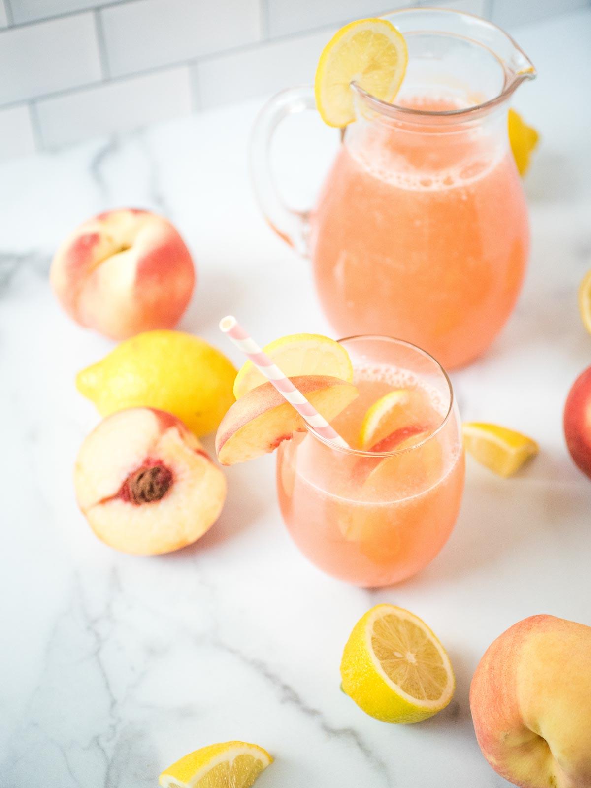 Come fare la Limonata alla Pesca Fatta in Casa 5 Un bicchiere di limonata alla pesca circondato da limoni e pesche con la grande brocca di limonata alla pesca sullo sfondo