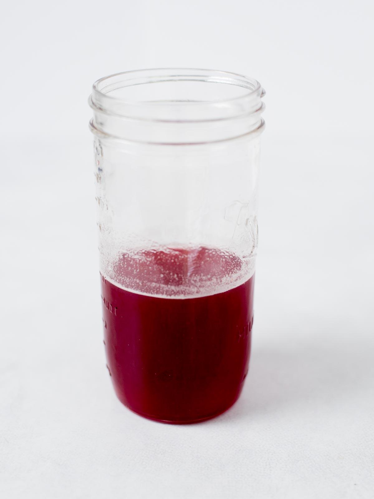Un barattolo di vetro trasparente contenente una piccola quantità di liquido rosso brillante, posato su una superficie bianca con uno sfondo bianco semplice.