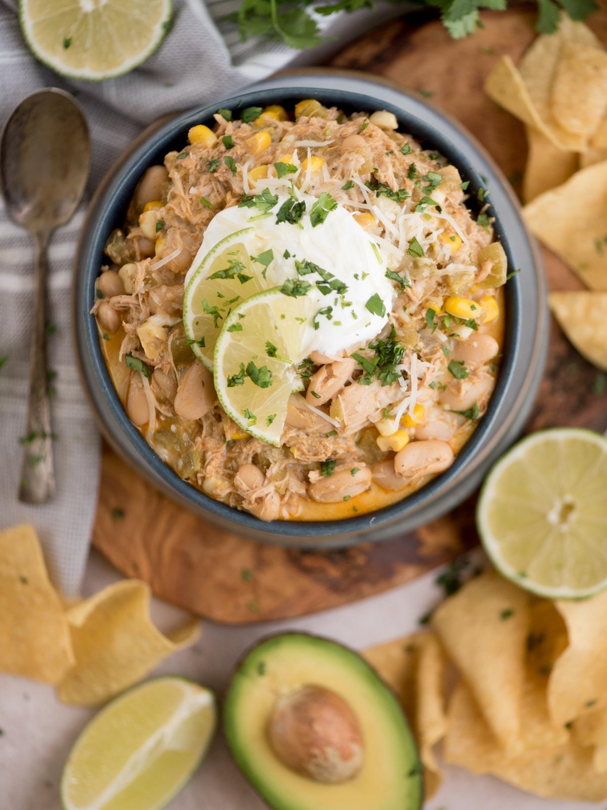 Chili di Pollo Bianco del Sud-Ovest 6 Una ciotola di chili bianco di pollo guarnito con panna acida, fette di lime e coriandolo. Circondato da metà avocado, lime, tortilla chips e un cucchiaio su un tagliere di legno.