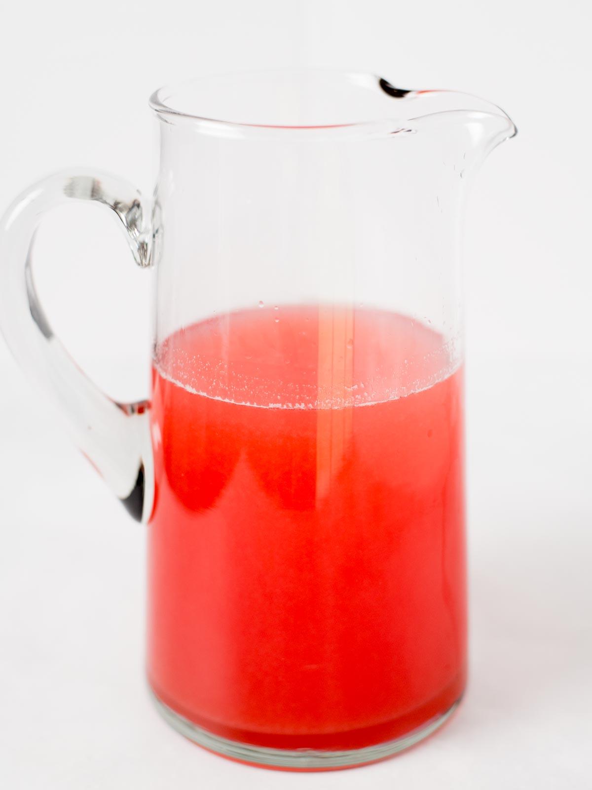 Una caraffa di vetro trasparente riempita a metà con liquido rosso brillante, posta su uno sfondo bianco semplice.