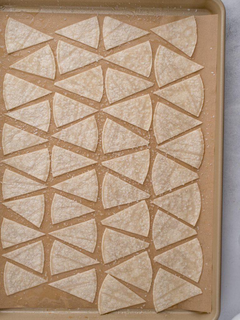 Teglia da forno con tortillas di mais tagliate a pezzi triangolari, condite con olio di avocado, salate e pronte per essere cotte.