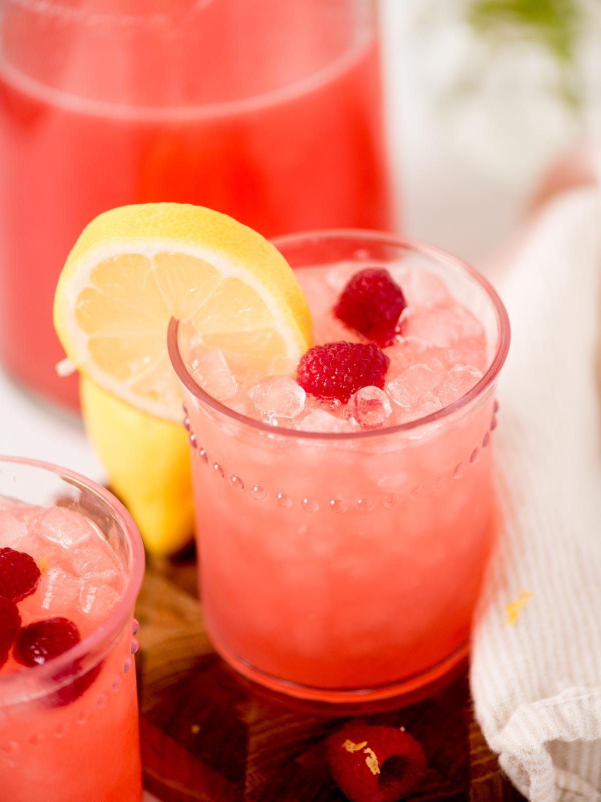 Un bicchiere di limonata rosa ai lamponi con ghiaccio tritato, guarnito con due lamponi e una fetta di limone. Una caraffa della stessa bevanda e un panno bianco sono sullo sfondo.