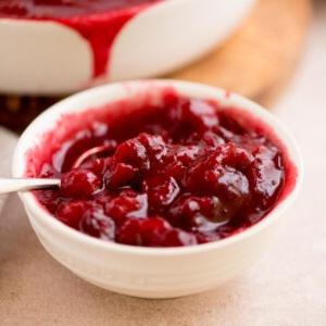 Salsa ai Mirtilli Rossi Interi 7 Una ciotola bianca piena di salsa di mirtilli rossi di colore rosso vibrante, contenente mirtilli interi e una consistenza lucida. Un cucchiaio è parzialmente immerso nella salsa. Lo sfondo mostra più salsa in un piatto bianco.