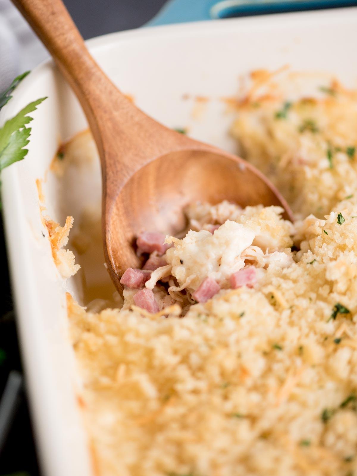 Teglia di pollo cordon bleu pronta da servire, con un cucchiaio da portata inserito. Attorno, prezzemolo fresco e un canovaccio da cucina.