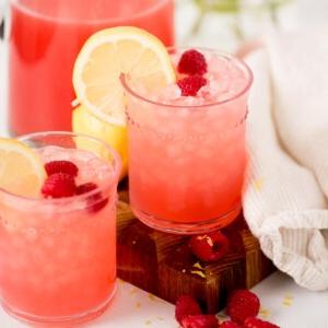 Due bicchieri di limonata rosa ai lamponi con ghiaccio, guarniti con fette di limone e lamponi, posati su un tagliere di legno accanto a un panno bianco e una caraffa della stessa bevanda. Lamponi freschi sono sparsi intorno.