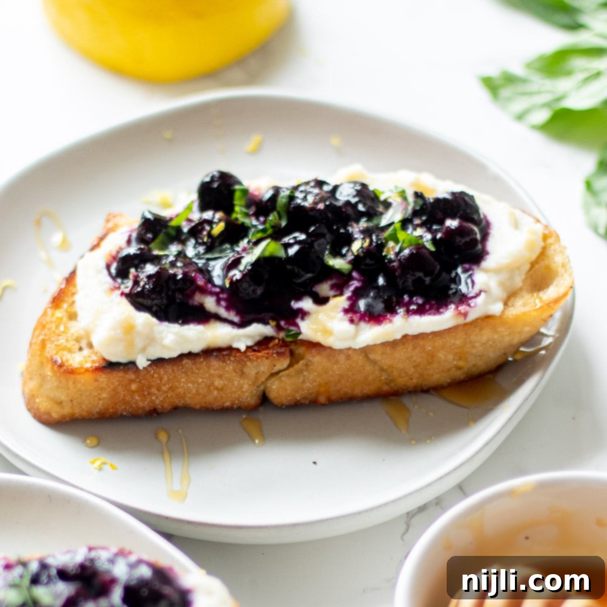 Creamy Whipped Ricotta and Blueberry Toast 2 ricotta blueberry toast on a plate with a fork and knife, garnished with basil