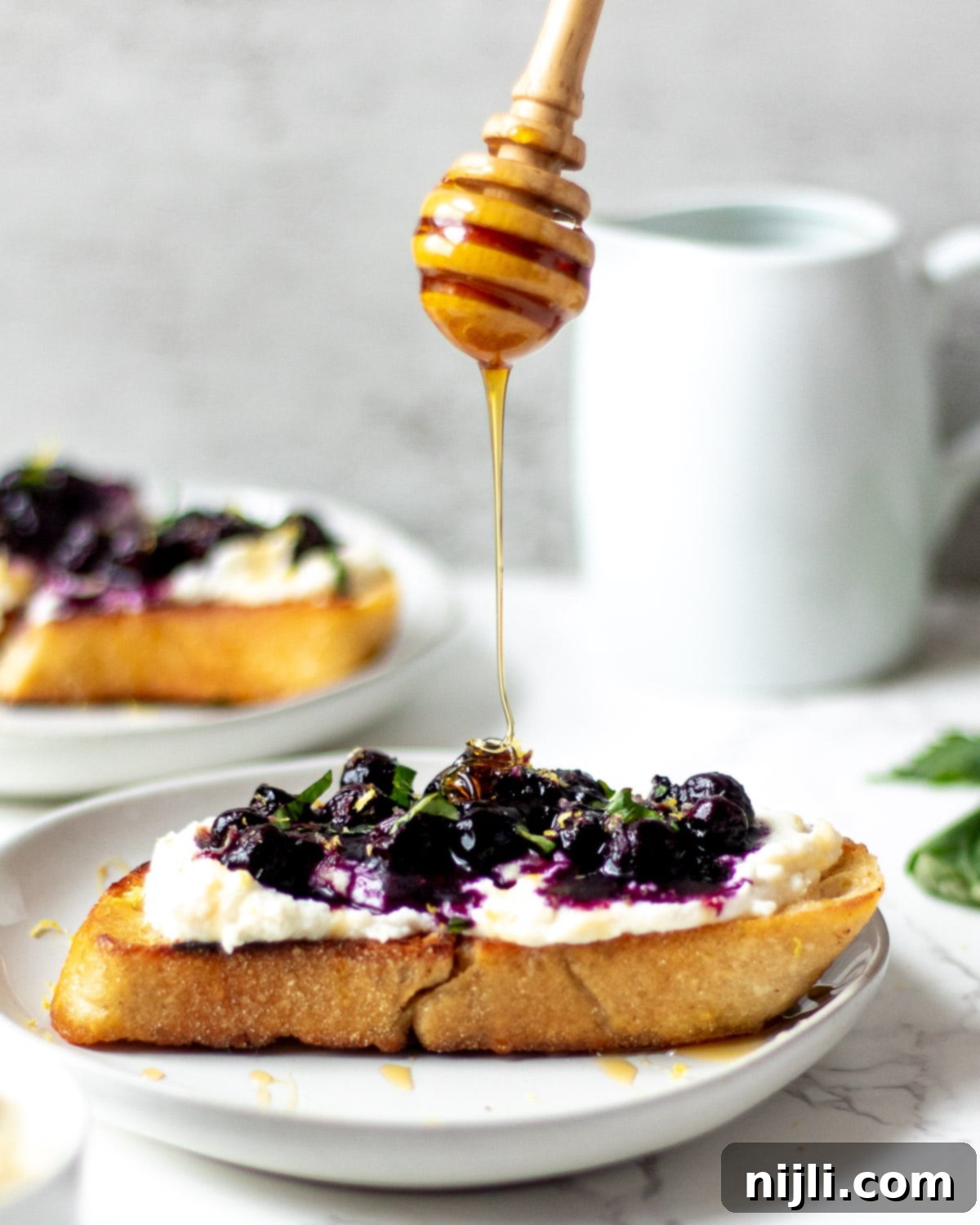 Creamy Whipped Ricotta and Blueberry Toast 3 honey drizzling over blueberry ricotta toast on a white plate