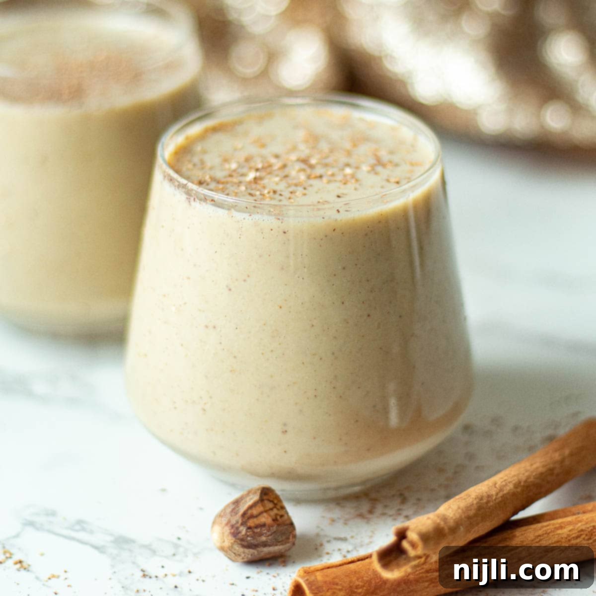 Spiced Oat Milk Cheer 2 Two glasses of creamy oat milk eggnog, garnished with freshly grated nutmeg, ready to be enjoyed during the holiday season.