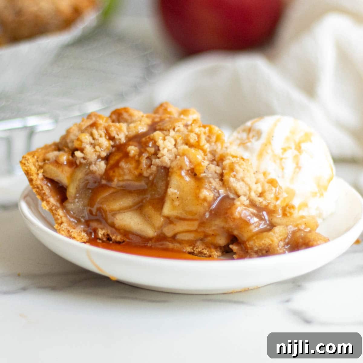 Irresistible Graham Cracker Apple Pie 2 A slice of apple pie in a graham cracker crust drizzled with caramel sauce and served with a scoop of vanilla ice cream on a white plate.