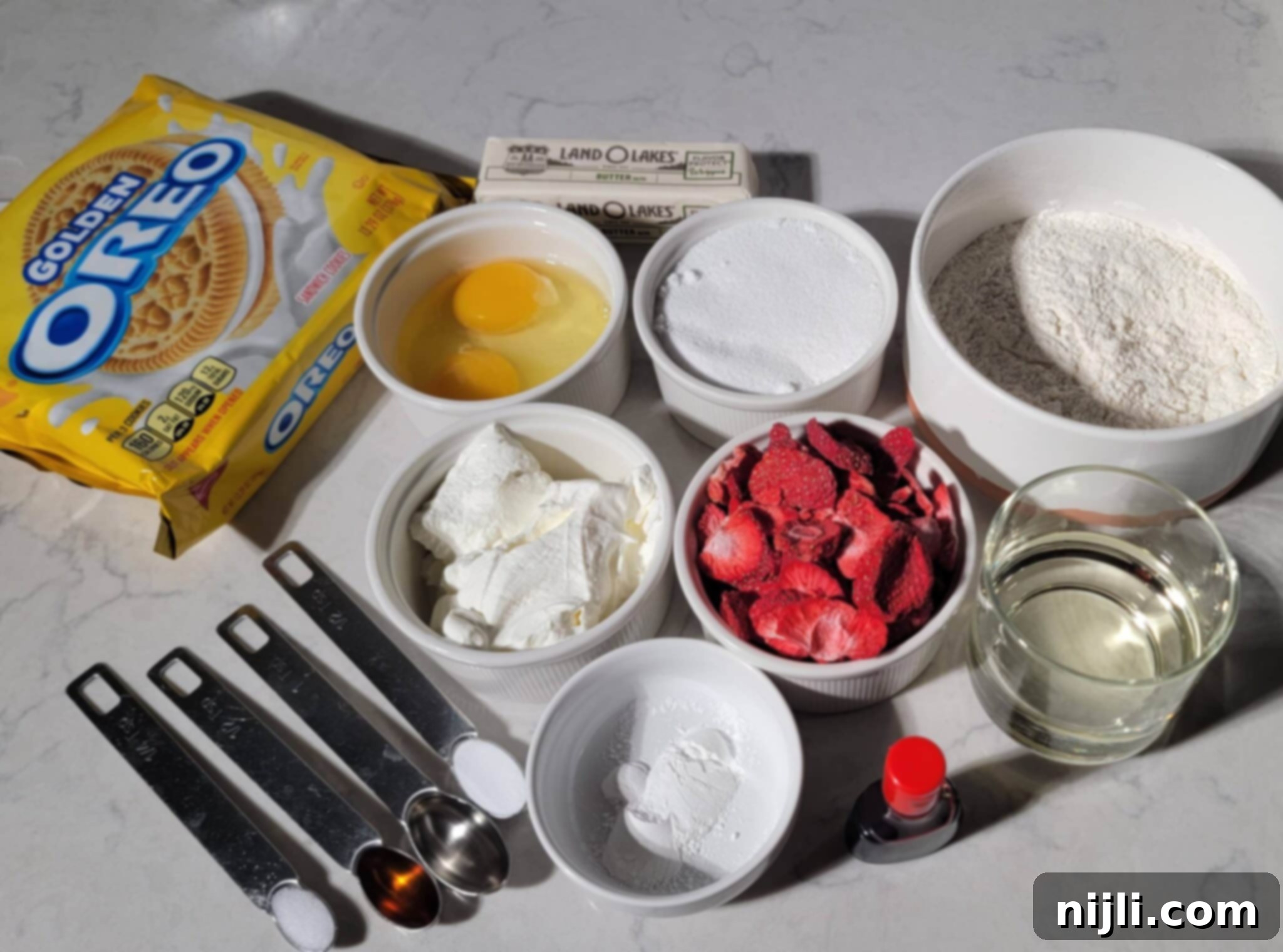 Strawberry Crunch Cupcakes 4 Ingredients for strawberry crunch cupcakes, featuring Golden Oreos, butter, eggs, flour, sugar, and freeze-dried strawberries on a marble counter.