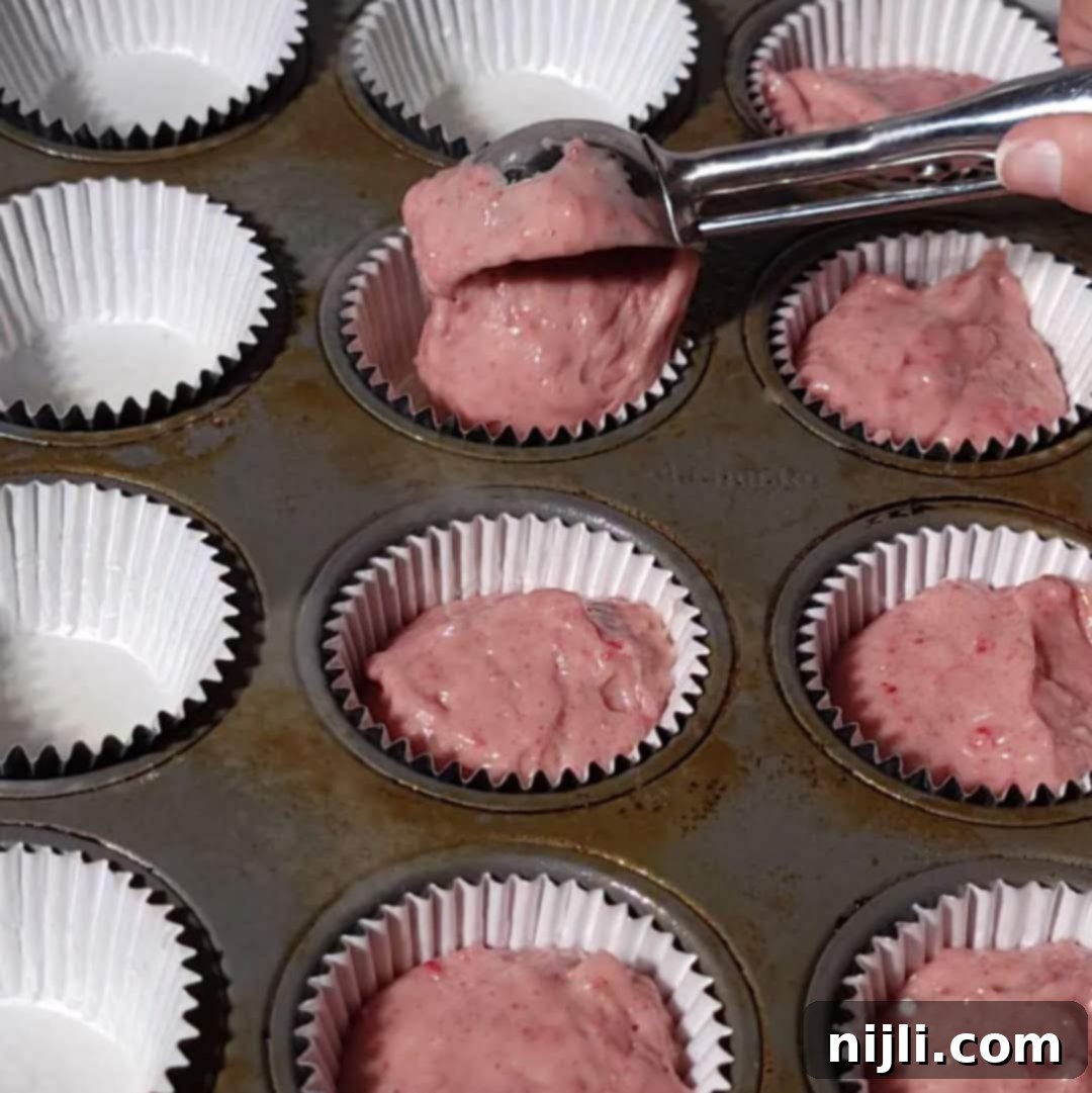 Strawberry Crunch Cupcakes 10 Portioning strawberry crunch cupcake batter into white liners in a muffin tin, preparation step for baking.