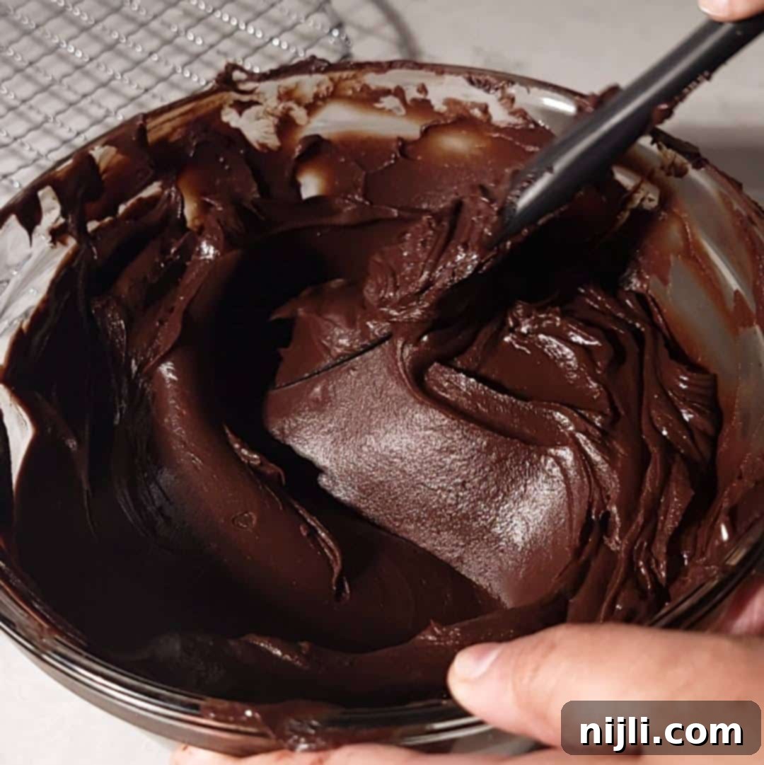 Matilda's Decadent Chocolate Cake 12 A glass bowl filled with shiny, dark chocolate fudge frosting, being stirred to perfection for the ultimate Matilda's Chocolate Cake topping.