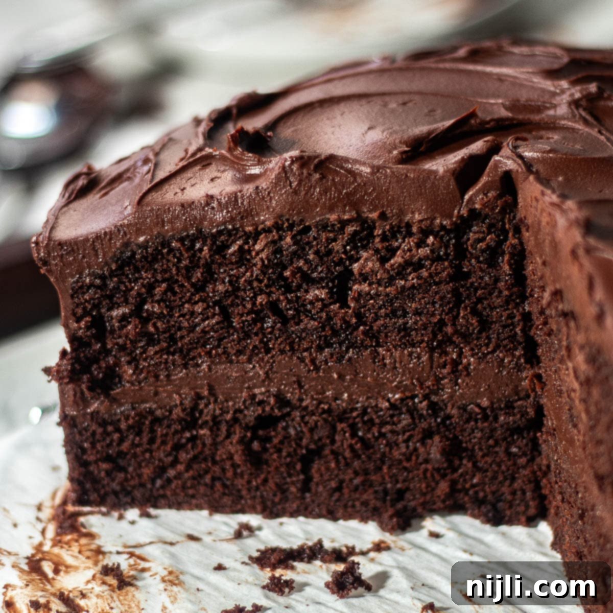 Matilda's Decadent Chocolate Cake 14 A perfectly cut slice of Matilda's Chocolate Cake, showcasing its moist layers and generous fudge frosting, resting against the remainder of the cake on a white marble surface, highlighting its rich texture.