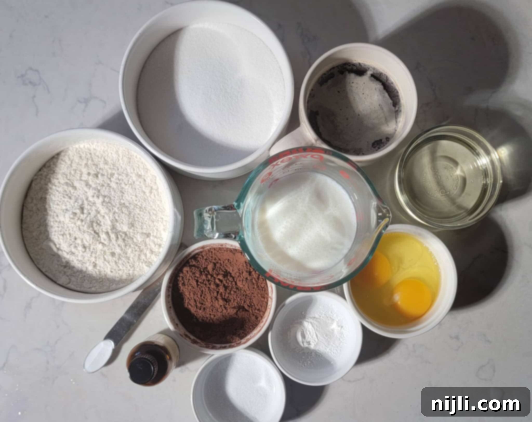 Matilda's Decadent Chocolate Cake 5 All the dry and wet ingredients neatly arranged on a white marble countertop for making Matilda's Chocolate Fudge Cake, including flour, cocoa powder, sugar, eggs, buttermilk, and hot coffee.