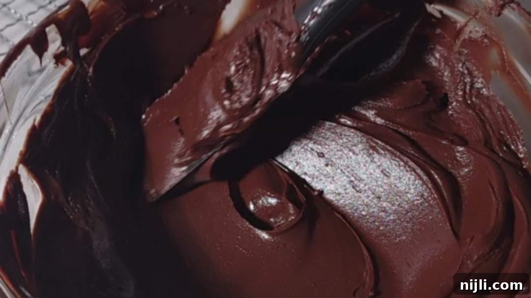 A spatula mixing thick chocolate fudge frosting in a bowl, illustrating the creamy consistency ideal for chocolate fudge frosting preparation.