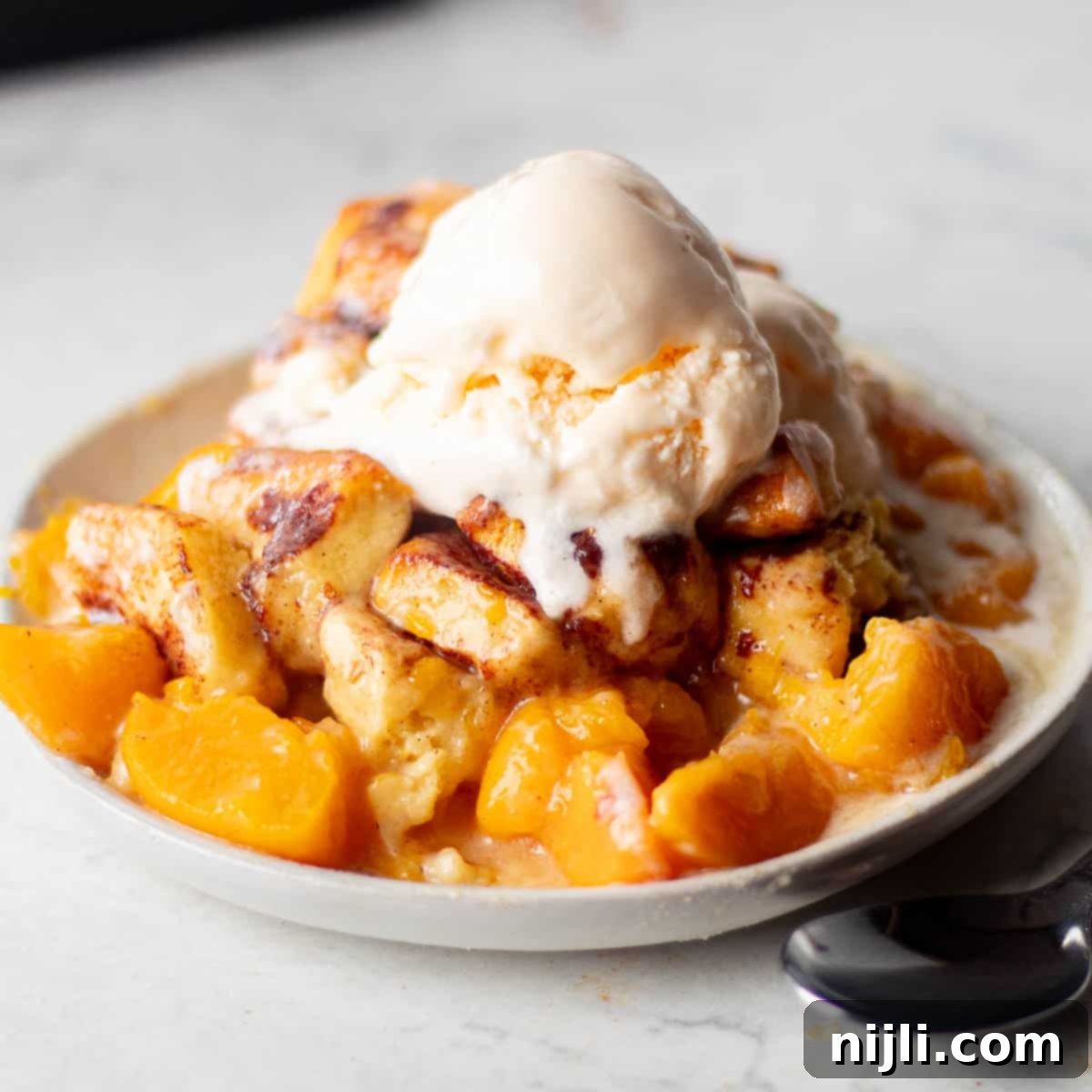 A serving of cinnamon rolls peach cobbler on a plate, topped with a scoop of vanilla ice cream.