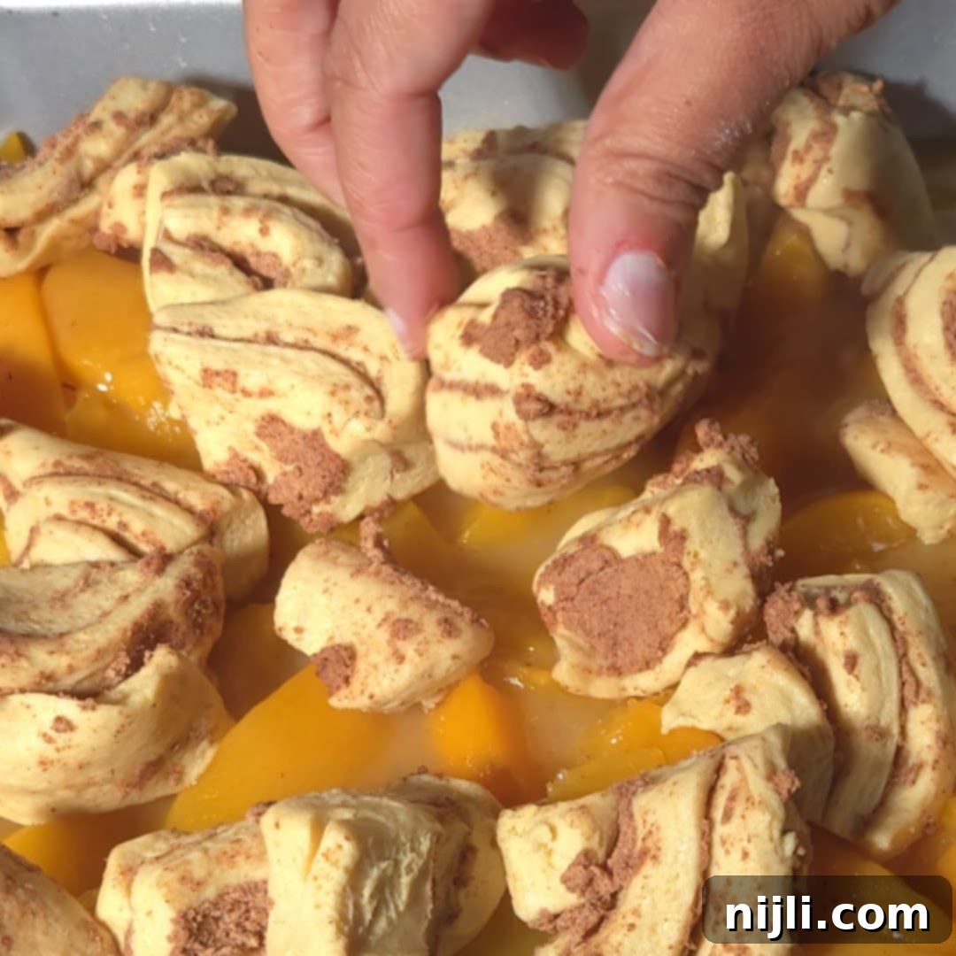 Hands placing cinnamon roll dough on top of peach slices in a baking pan, showing the preparation process of cinnamon rolls peach cobbler.