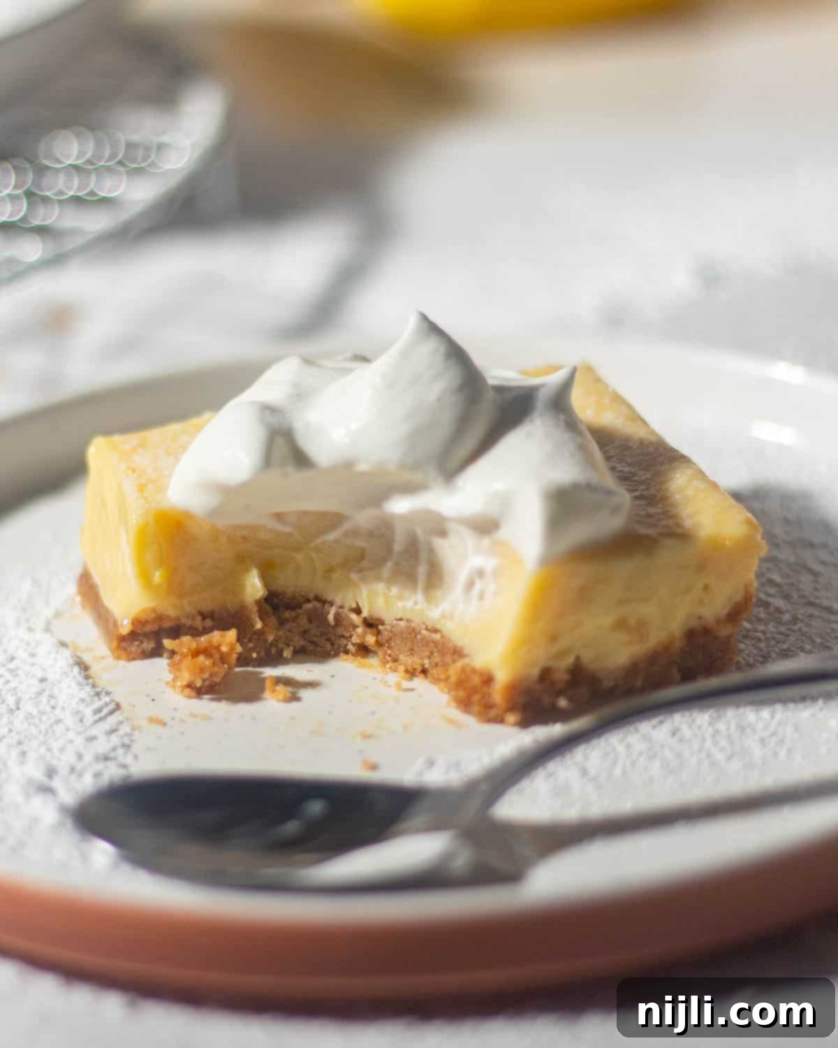 Golden Lemon Delight with Graham Crumb Perfection 3 A partially eaten lemon bar with a graham cracker crust and whipped cream on top, served on a white plate with a spoon next to it, showing the creamy lemon filling.