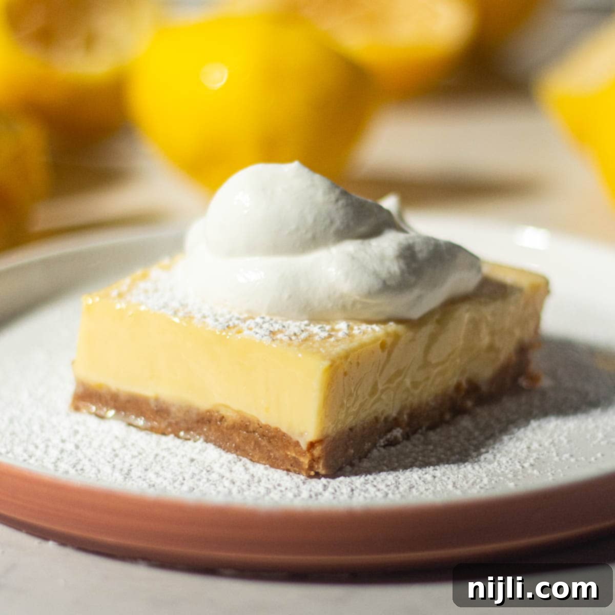 Golden Lemon Delight with Graham Crumb Perfection 16 A single lemon bar with a graham cracker crust on a white plate, topped with whipped cream and lightly dusted with powdered sugar, surrounded by halved lemons.