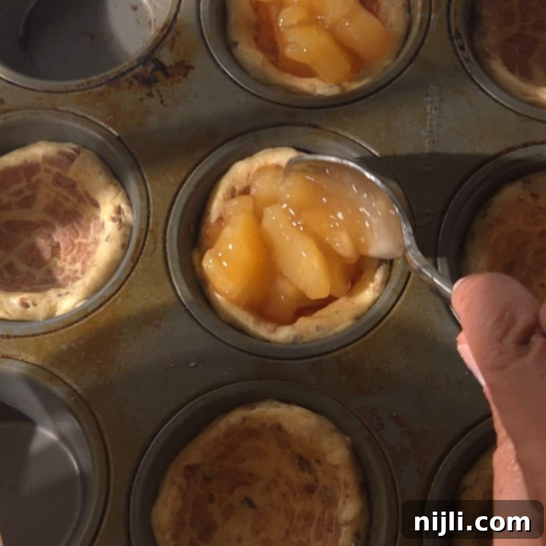 Cinnamon Swirl Apple Pies 8 A hand spooning apple pie filling into cinnamon roll dough placed in a muffin tin. This shows a step in making cinnamon roll apple pies.