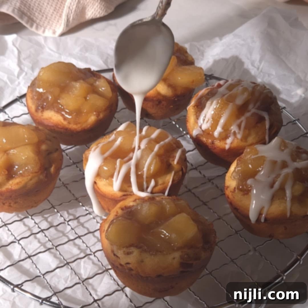 Cinnamon Swirl Apple Pies 9 Freshly baked cinnamon roll apple pies on a wire rack with icing being drizzled on top using a spoon. The apple pie filling is visible, creating a delicious and easy dessert.