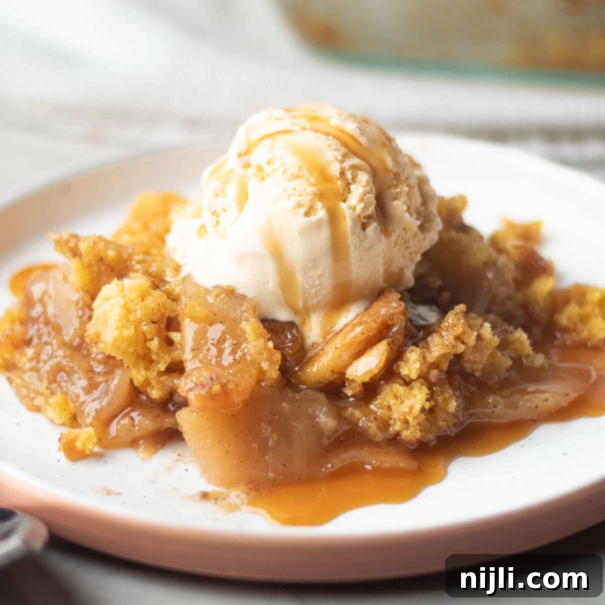 Effortless Cake Mix Apple Cobbler 2 A plate of apple cobbler with yellow cake mix, topped with vanilla ice cream and drizzled with caramel sauce. The golden, crumbly topping covers soft, cinnamon-spiced apple slices.