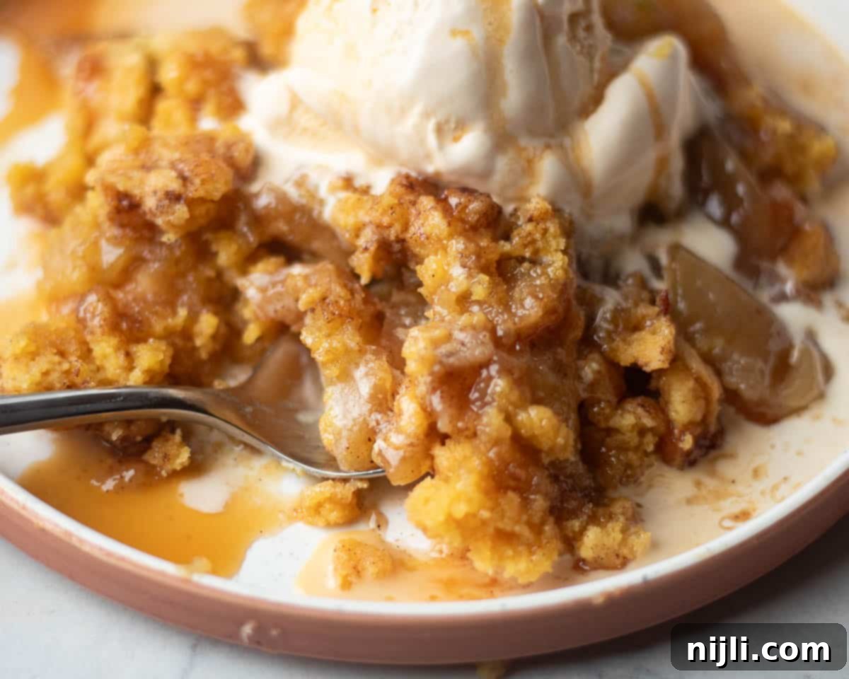 A fork digging into a serving of apple cobbler with yellow cake mix, topped with melting vanilla ice cream. The dish has a crispy, buttery topping over tender apples.