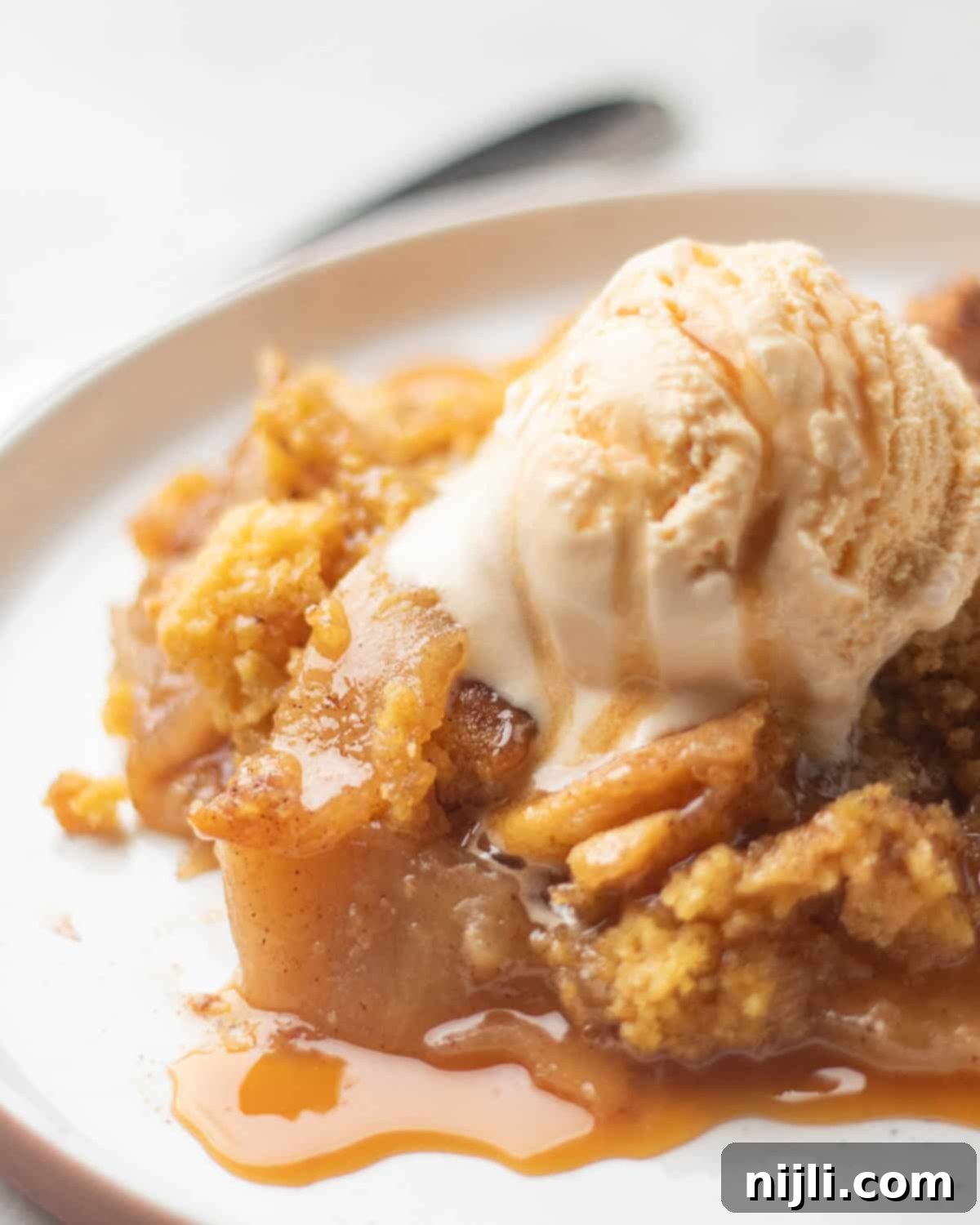 Effortless Cake Mix Apple Cobbler 3 A golden-brown apple cobbler made with canned apples and yellow cake mix, topped with a scoop of vanilla ice cream and drizzled with caramel sauce.