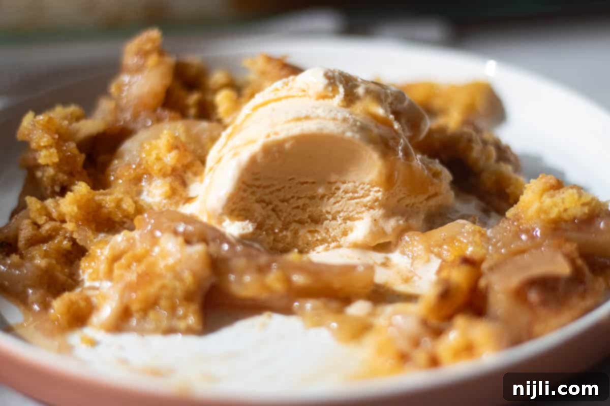 Effortless Cake Mix Apple Cobbler 5 A close-up of apple cobbler made with yellow cake mix, topped with a scoop of vanilla ice cream and drizzled with caramel sauce. The cobbler has a golden, crumbly crust with tender apple slices.