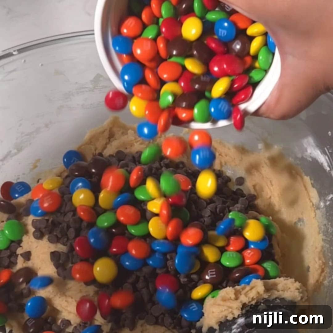 M&M Burst Chocolate Chip Goodness 12 A hand pouring a bowl of vibrant M&M candies and mini chocolate chips into the cookie dough, mixing for an even distribution. This step adds a fun, colorful twist to the M&M cookie recipe, ensuring every bite is filled with chocolate and candy pieces.