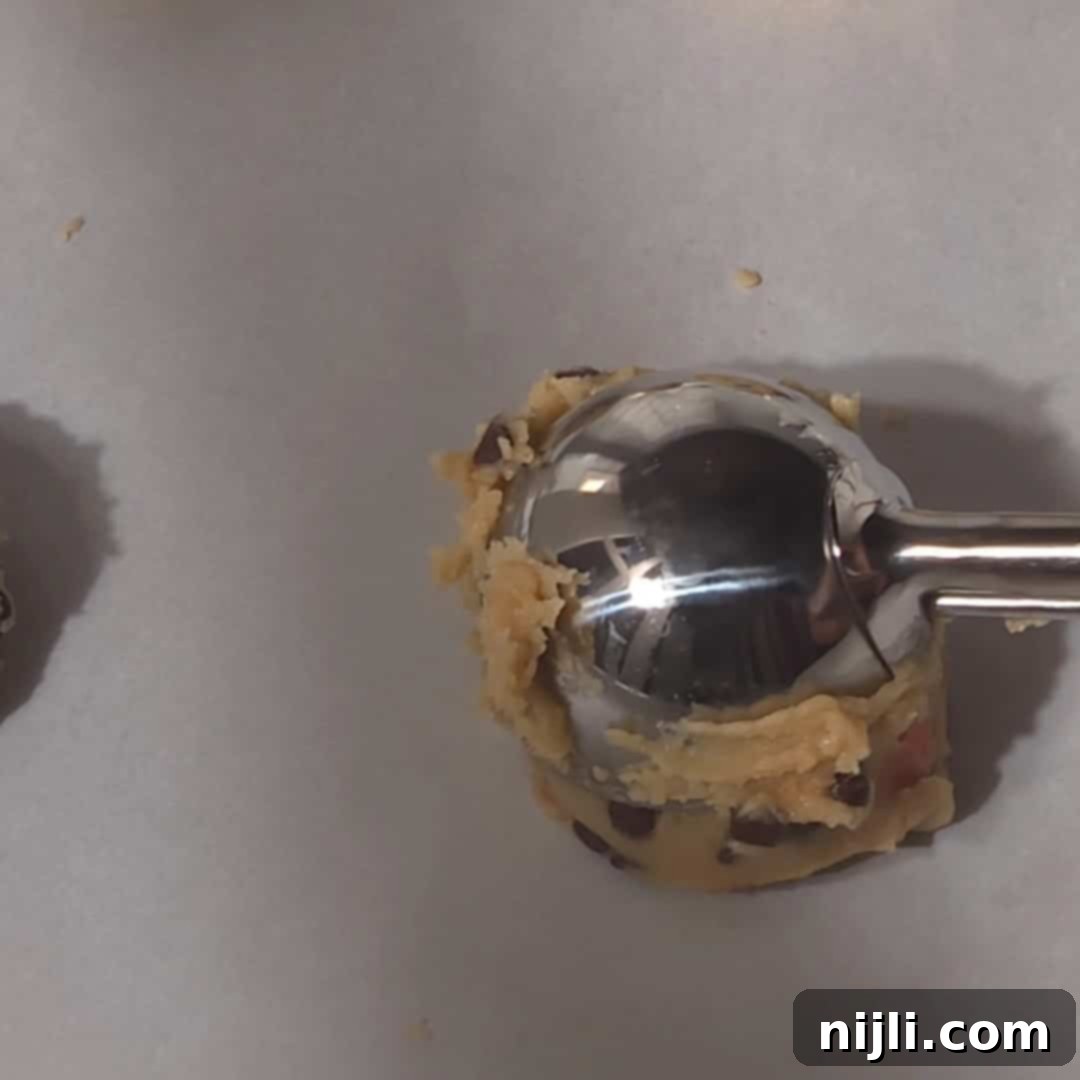 M&M Burst Chocolate Chip Goodness 13 A cookie scoop portioning out rounds of M&M cookie dough onto a baking sheet, forming evenly sized dough balls. This step ensures consistent cookie sizes, helping them bake uniformly in the oven.